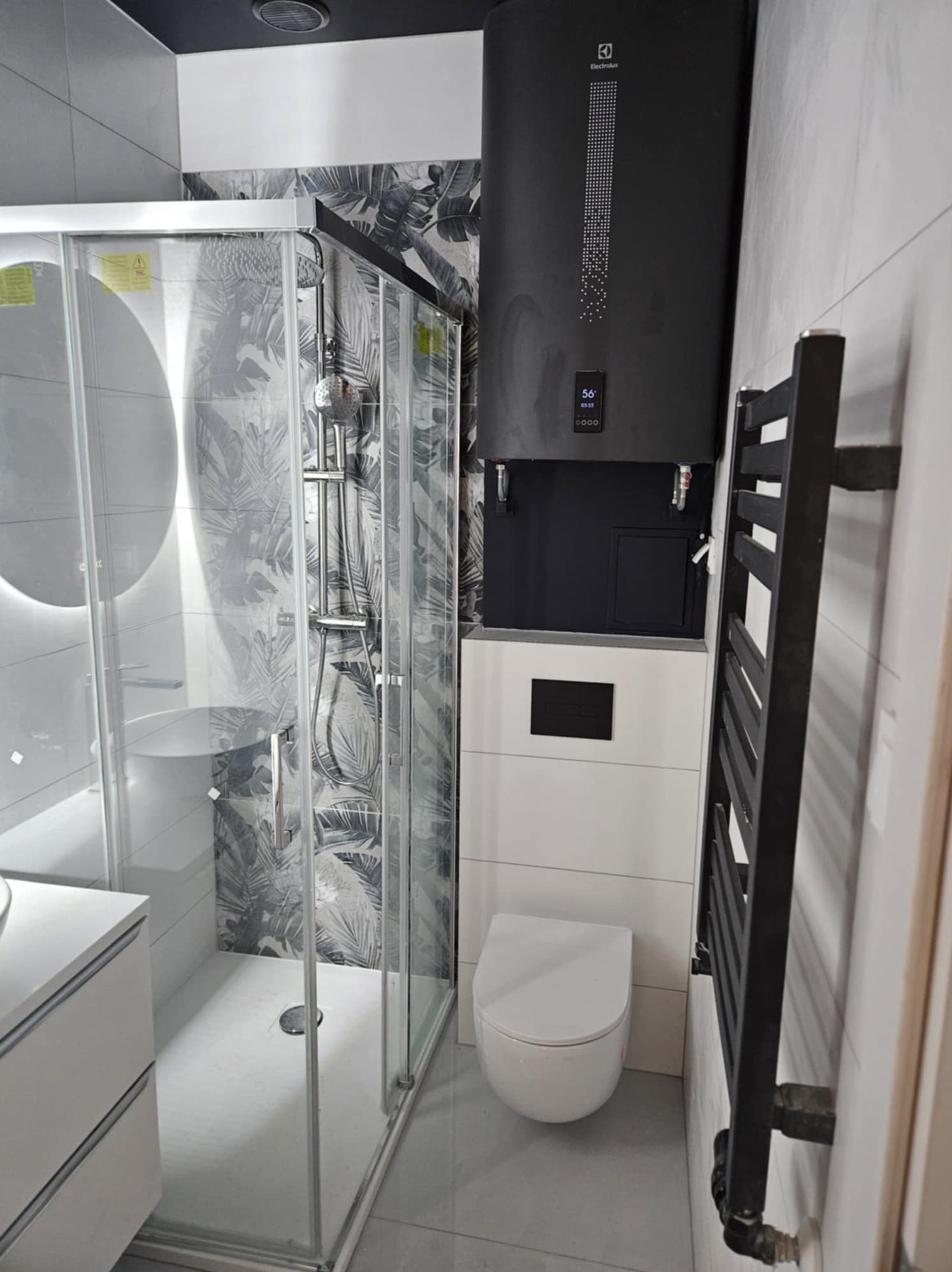 Nowoczesna łazienka w odcieniach szarości i bieli z czarnymi akcentami, kabina prysznicowa z wzorem liści na ścianie, wiszący bojler Electrolux i grzejnik drabinkowy.