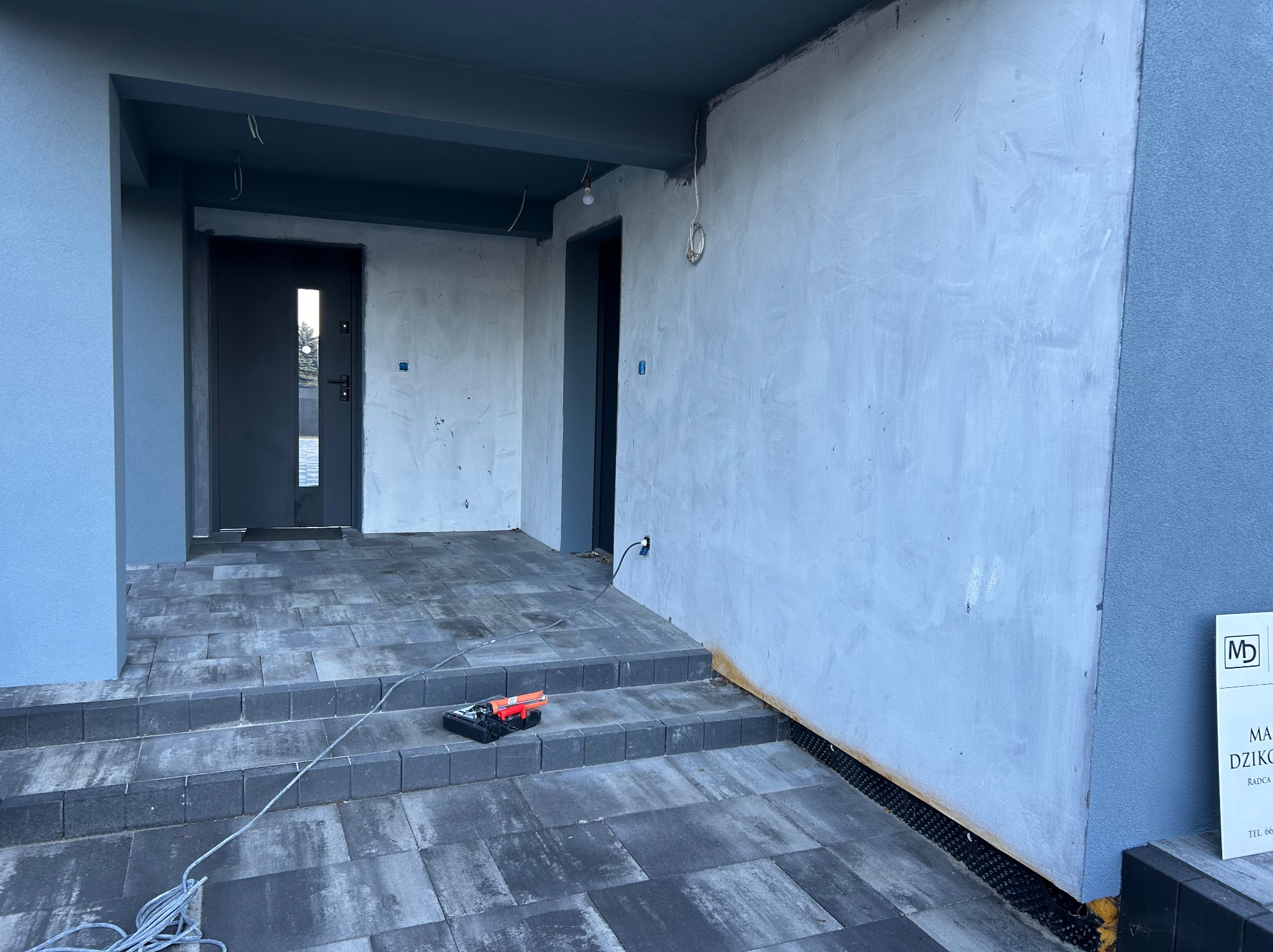 Surowy stan werandy z szarymi płytami chodnikowymi, ściany w kolorze szarym, widoczne narzędzia glazurnicze i kable, w tle drzwi wejściowe.