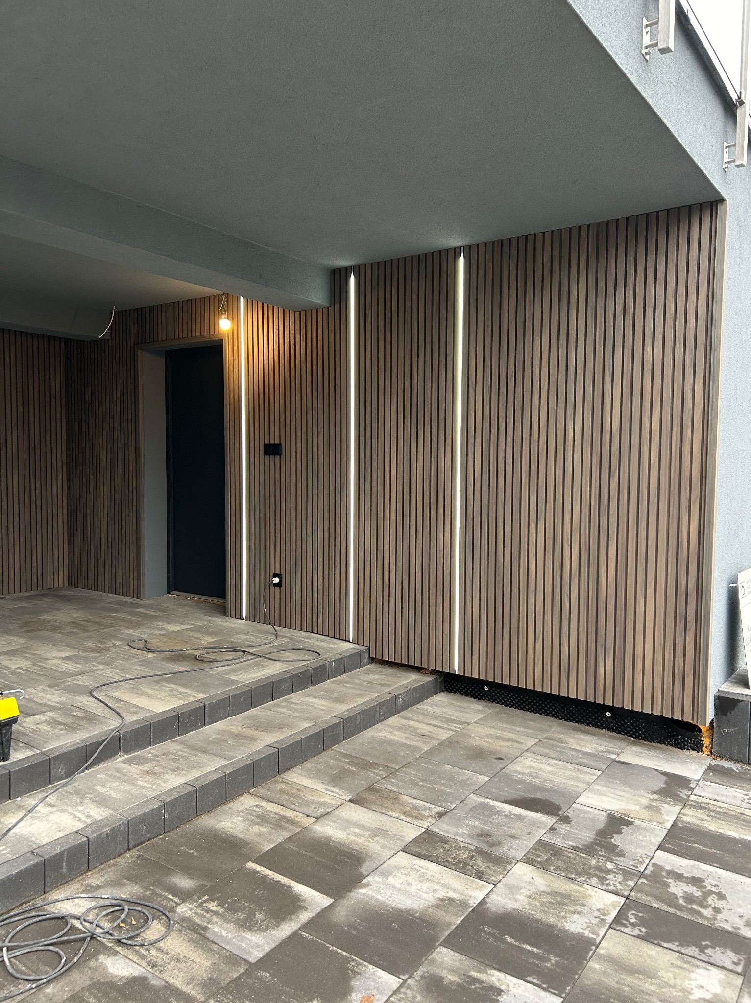 Nowoczesna elewacja z drewnianych paneli pionowych z wbudowanym oświetleniem LED, szare płytki chodnikowe i schody, czarne drzwi wejściowe.