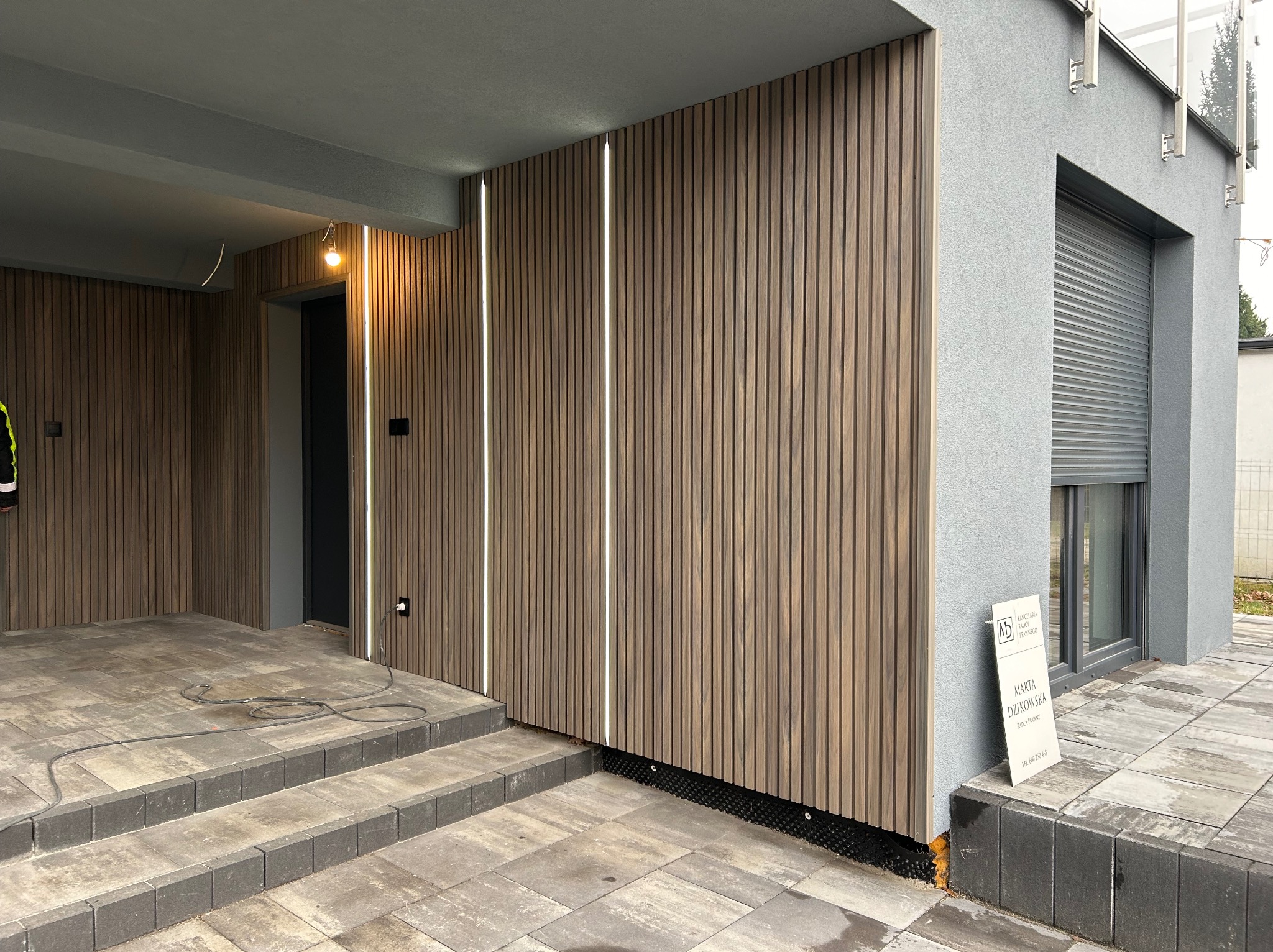 Nowoczesna elewacja budynku z drewnianymi panelami pionowymi i wbudowanym oświetleniem LED, widoczne schody wejściowe z kostki brukowej i fragment okna z roletą zewnętrzną.