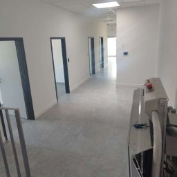 GrotClean - Długi, jasny korytarz biurowy z szarymi drzwiami i podłogą z płytek, widok z perspektywy urządzenia czyszczącego z czerwoną gałką, na końcu korytarza widoczne okno z naturalnym światłem.