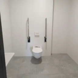 Biała toaleta podwieszana z chromowanymi uchwytami dla niepełnosprawnych, zamontowana w minimalistycznej łazience z szarą podłogą i białymi ścianami.