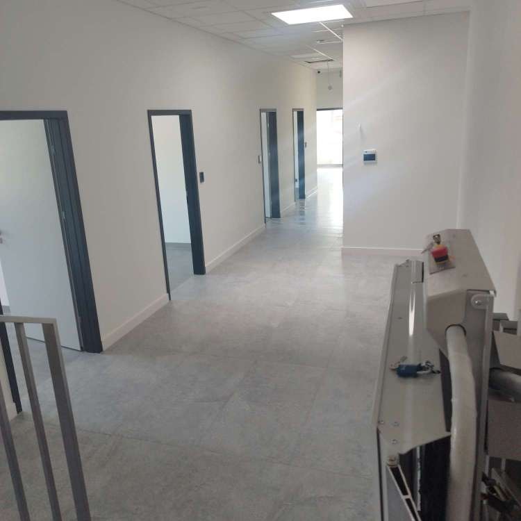 Długi, jasny korytarz biurowy z szarymi drzwiami i podłogą z płytek, widok z perspektywy urządzenia czyszczącego z czerwoną gałką, na końcu korytarza widoczne okno z naturalnym światłem.