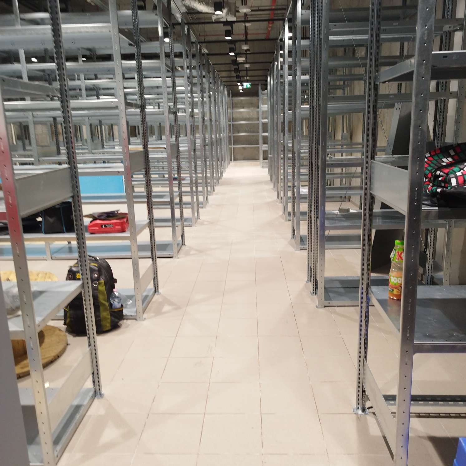 Długi korytarz z metalowymi regałami w magazynie, niektóre półki puste, inne z torbami i butelką soku; jasne płytki na podłodze, widoczne oświetlenie sufitowe.