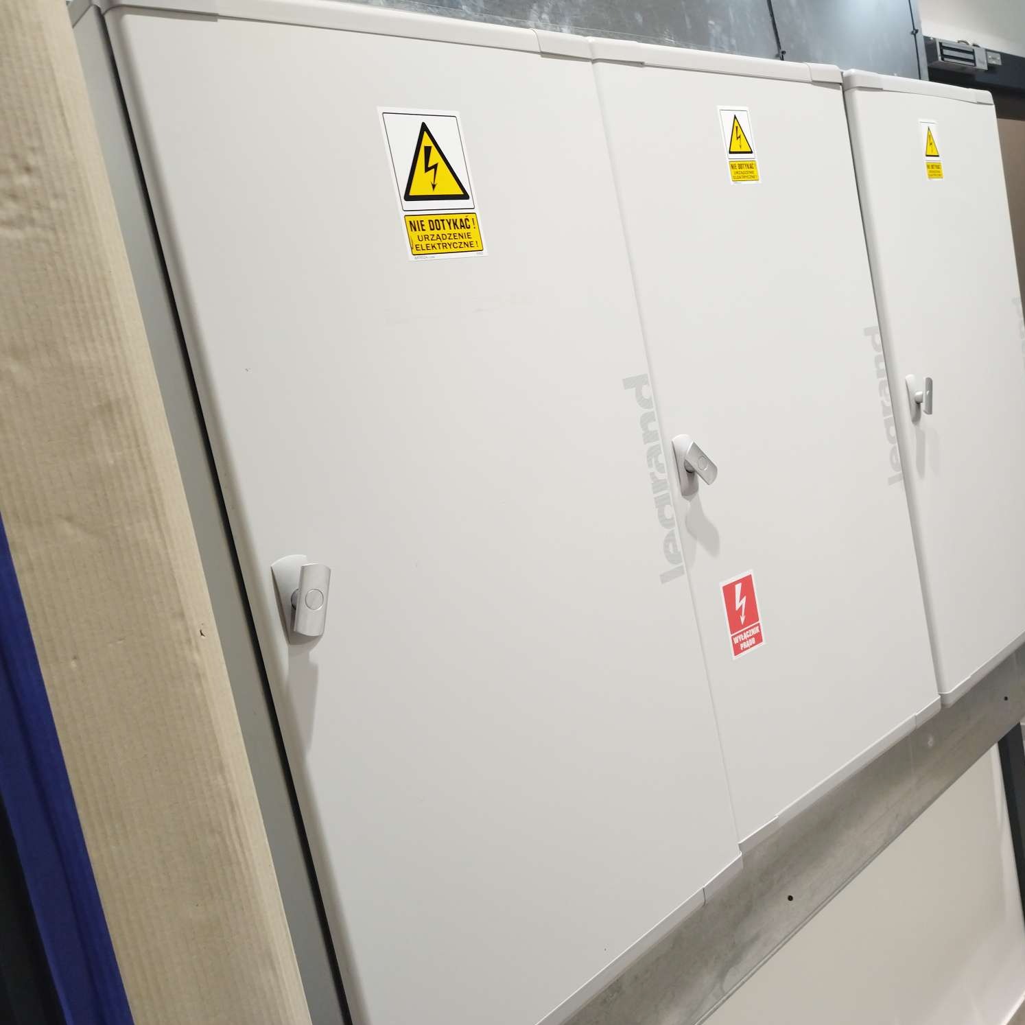 Białe szafy elektryczne marki Legrand z ostrzeżeniami o wysokim napięciu i wyłącznikiem prądu, widok z boku.