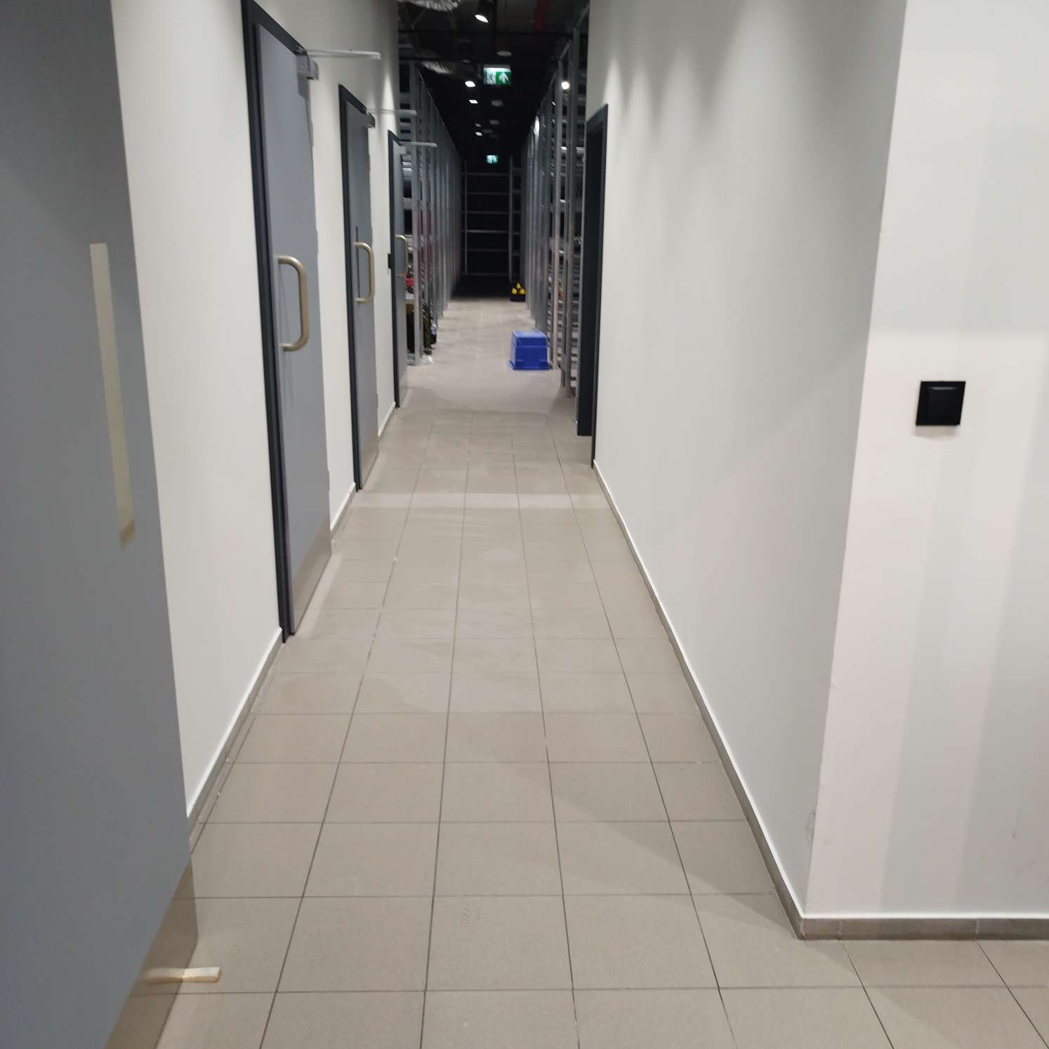 Długi, pusty korytarz biurowy z szarą podłogą z płytek, białymi ścianami i rzędem szarych drzwi po lewej stronie, z niebieskim stołkiem w oddali, sugerujący świeżo posprzątane pomieszczenie.