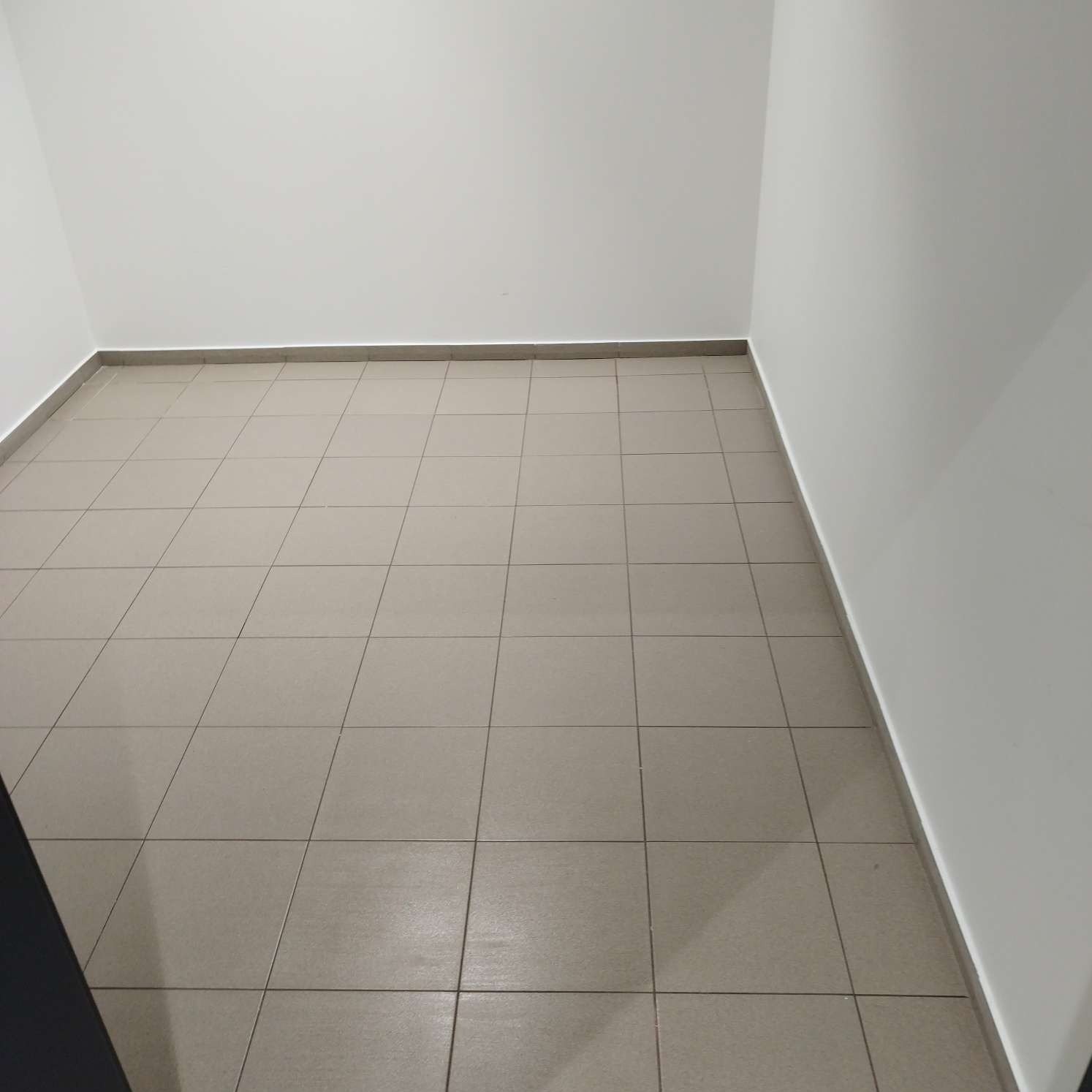Puste pomieszczenie z beżowymi płytkami na podłodze i białymi ścianami, widoczne szare fugi i listwy przypodłogowe.