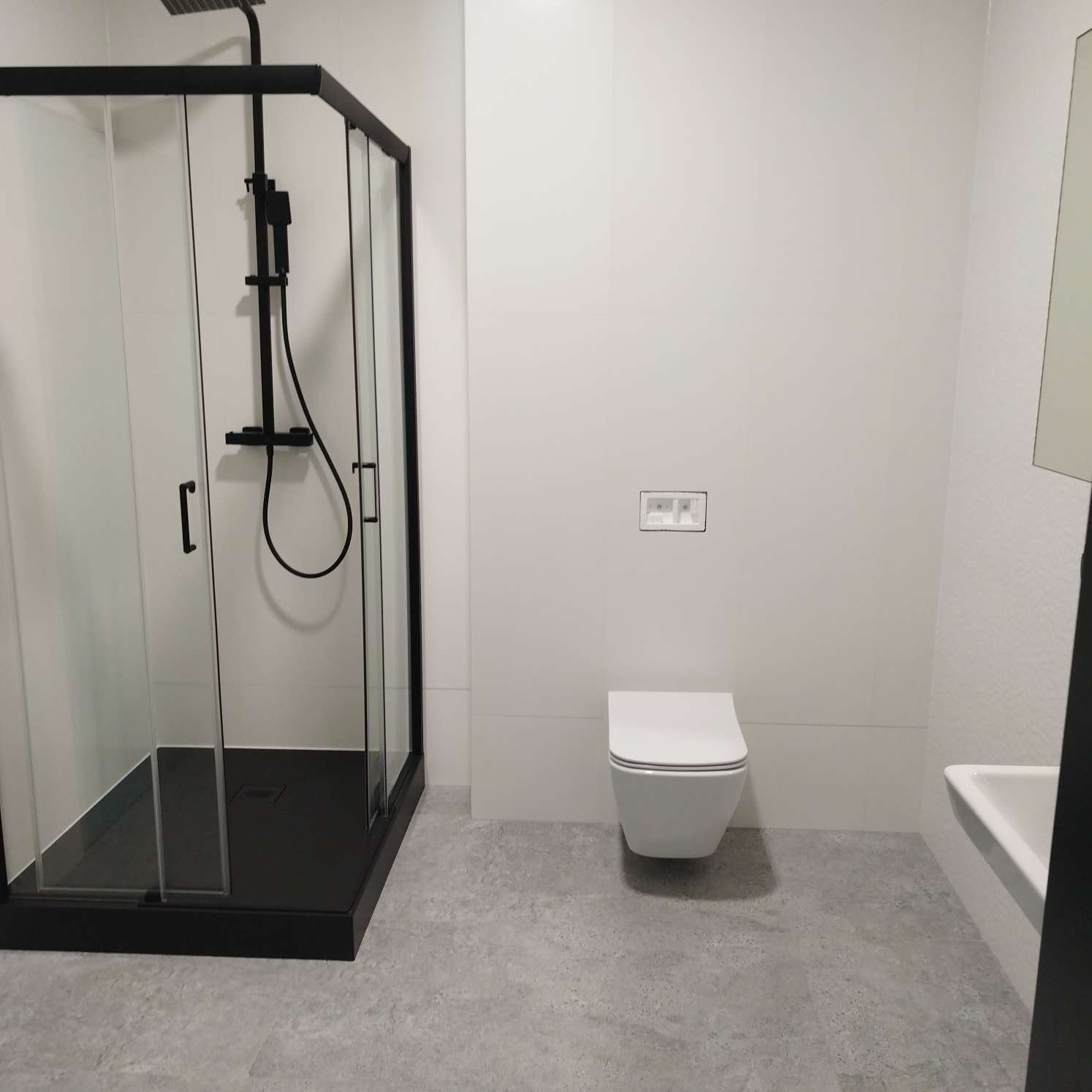 Minimalistyczna łazienka z czarną kabiną prysznicową typu walk-in, podwieszaną toaletą i umywalką, wykończona w bieli i szarości.