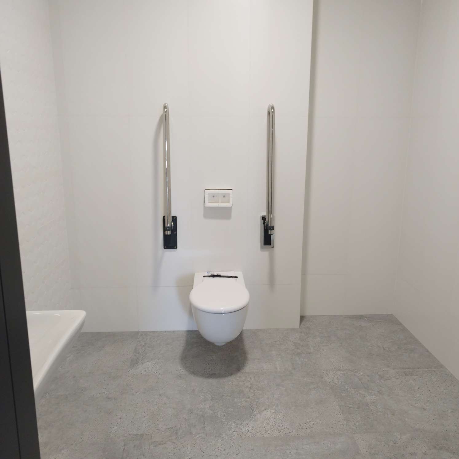 Biała toaleta podwieszana z uchwytami dla niepełnosprawnych na tle białych płytek, szara podłoga, umywalka z lewej strony kadru.