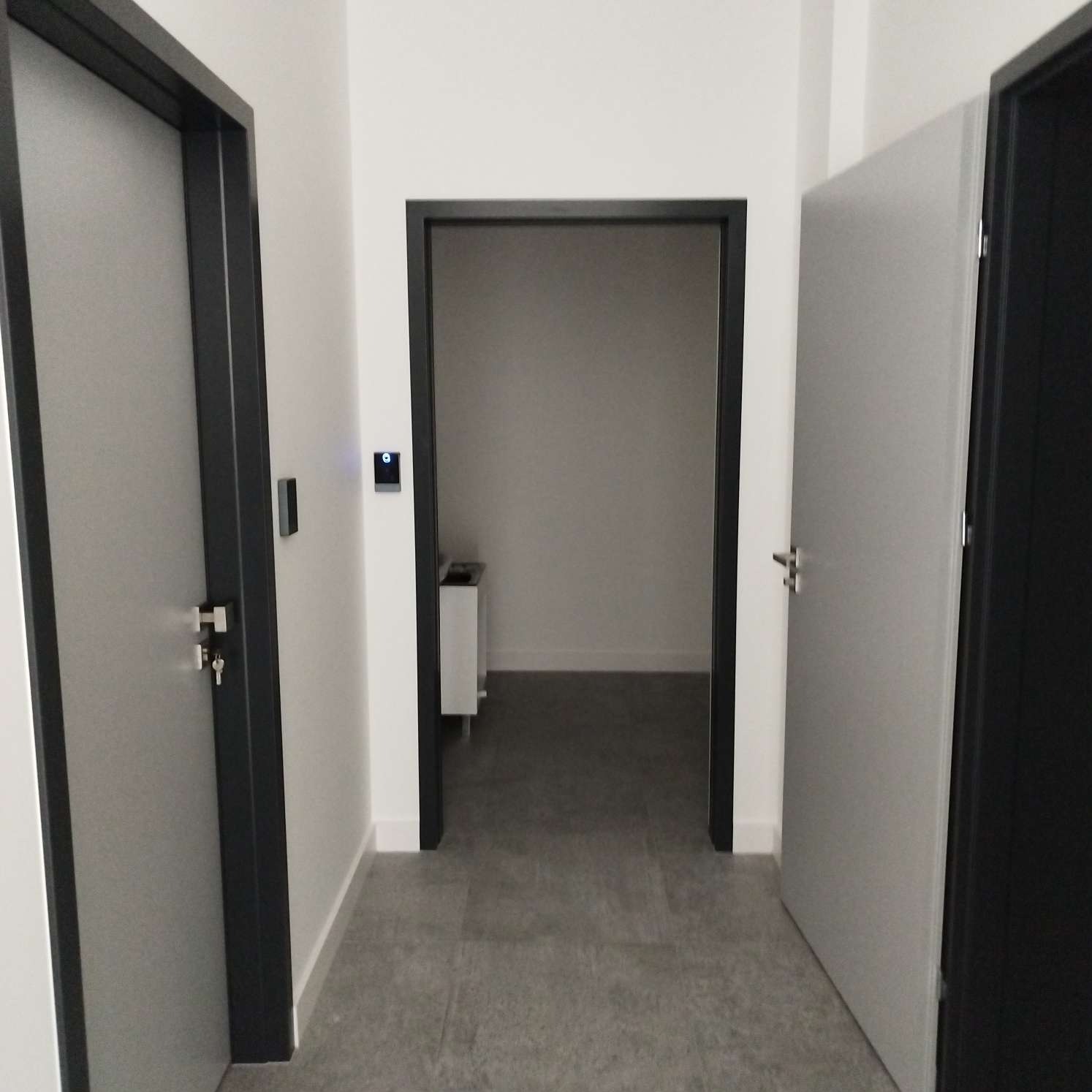 Wnętrze korytarza z trzema drzwiami, szarą podłogą i białymi ścianami, widok na puste pomieszczenie w tle.