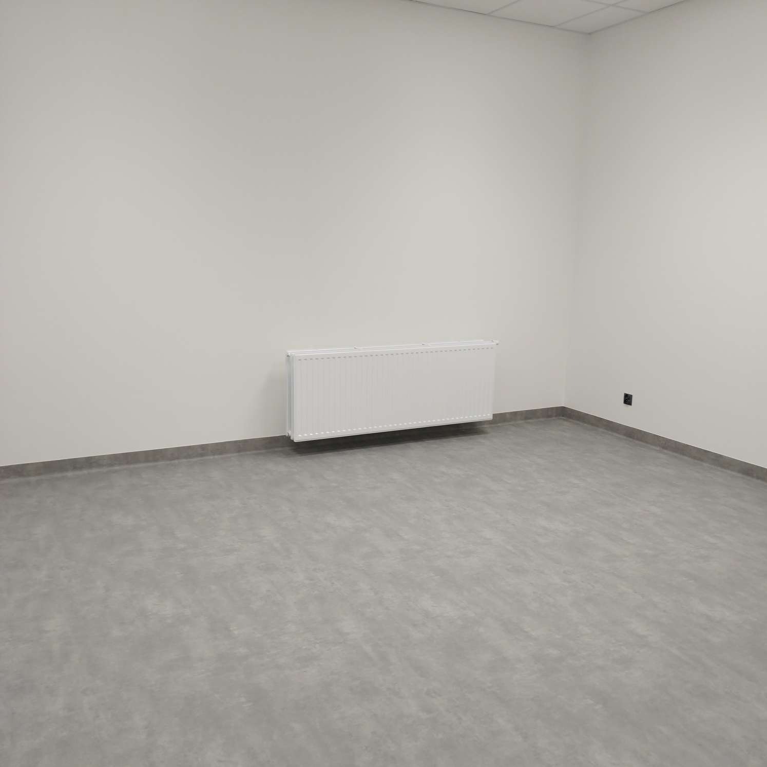 Puste, jasne pomieszczenie z szarą podłogą, białymi ścianami i grzejnikiem ściennym. Widoczny niski cokół wzdłuż ścian.