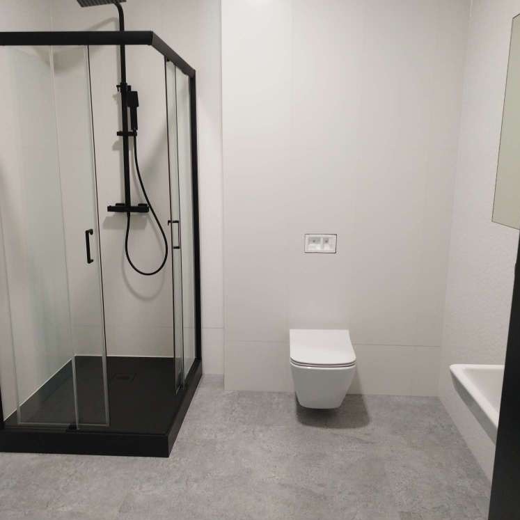 Minimalistyczna łazienka z czarną kabiną prysznicową typu walk-in, wiszącą toaletą i szarą podłogą z płytek imitujących beton.