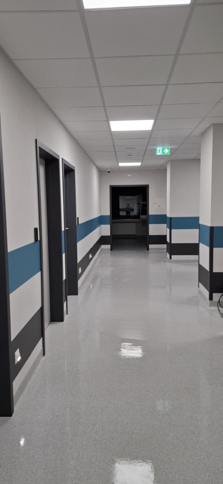 Wypolerowany, jasnoszary korytarz biurowy z czarnymi futrynami drzwi i pasami w kolorze niebieskim, białym i czarnym na ścianach; widoczny znak ewakuacyjny i osoba w oddali.