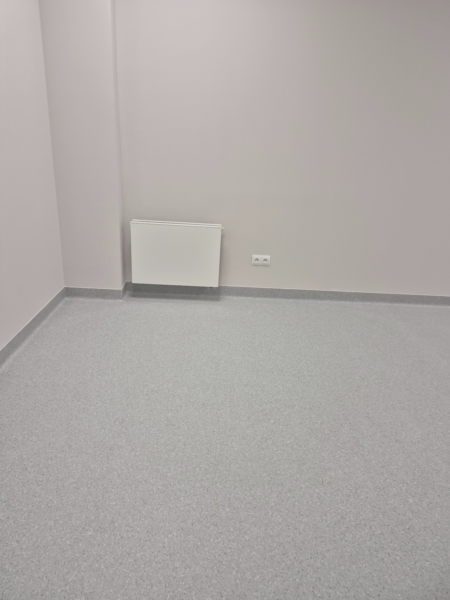 Puste, jasne pomieszczenie z szarą podłogą z tworzywa, grzejnikiem ściennym i dwoma gniazdkami elektrycznymi.