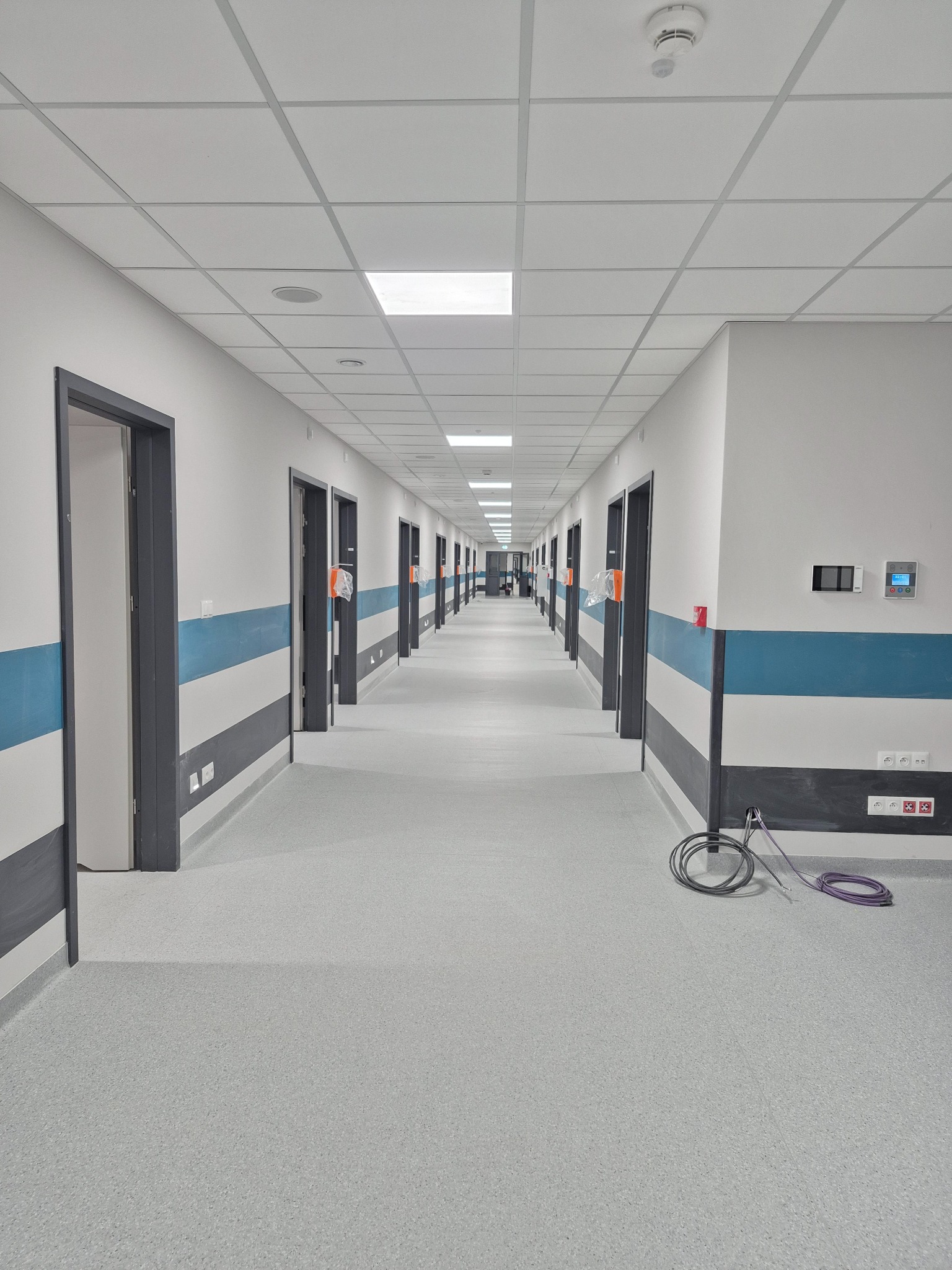 Długi korytarz w nowym budynku z szarymi drzwiami, niebieskimi pasami na ścianach i szarą podłogą, widoczne kable na podłodze.