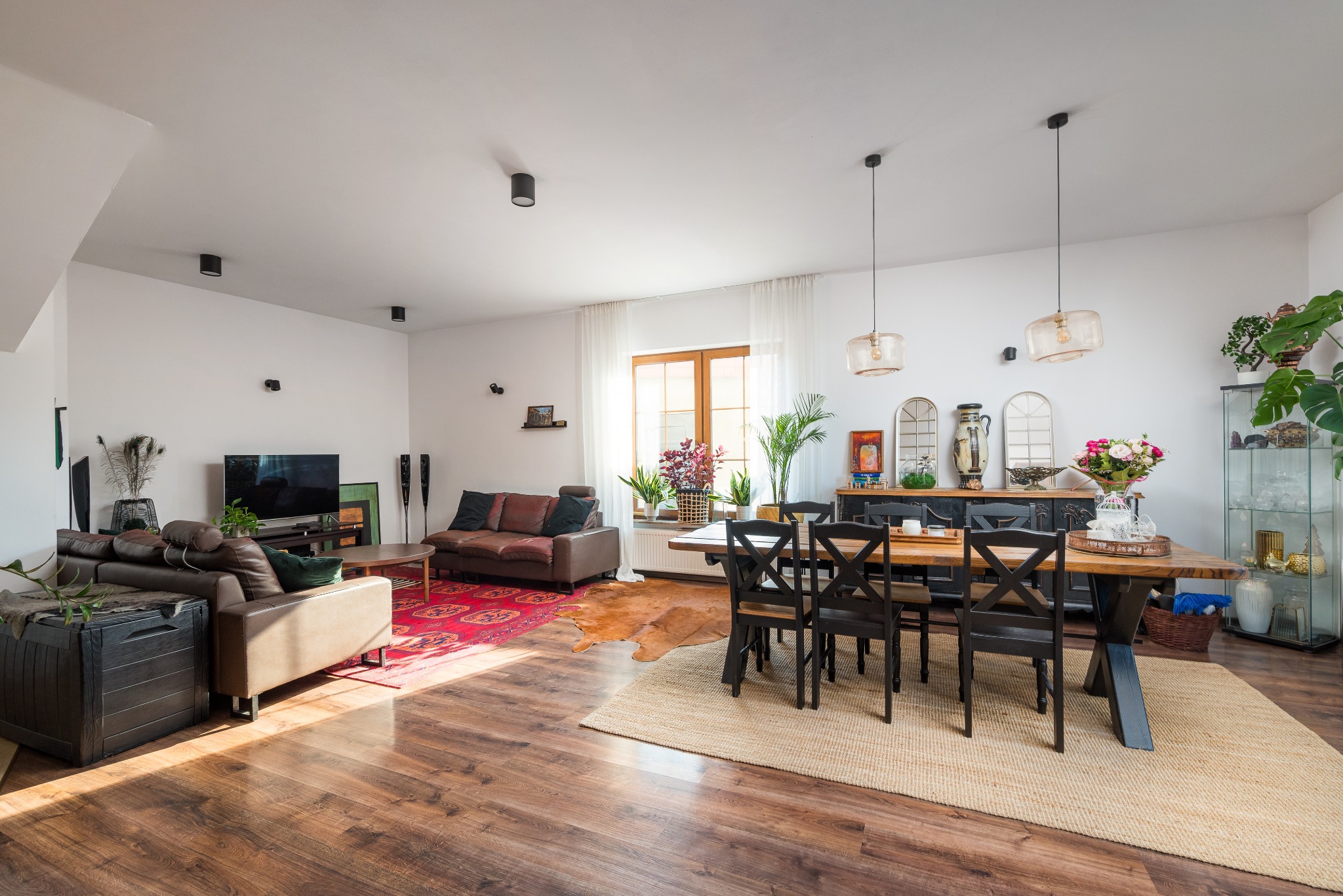 Przestronny salon z drewnianą podłogą, skórzanymi sofami, stołem jadalnym z czarnymi krzesłami i dekoracyjnymi elementami, oświetlony naturalnym światłem wpadającym przez okno.