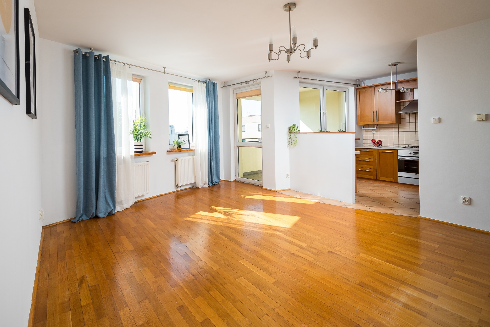 Jasne, przestronne wnętrze mieszkania z drewnianą podłogą, oknami z niebieskimi zasłonami, widokiem na balkon i kuchnię z drewnianymi szafkami.