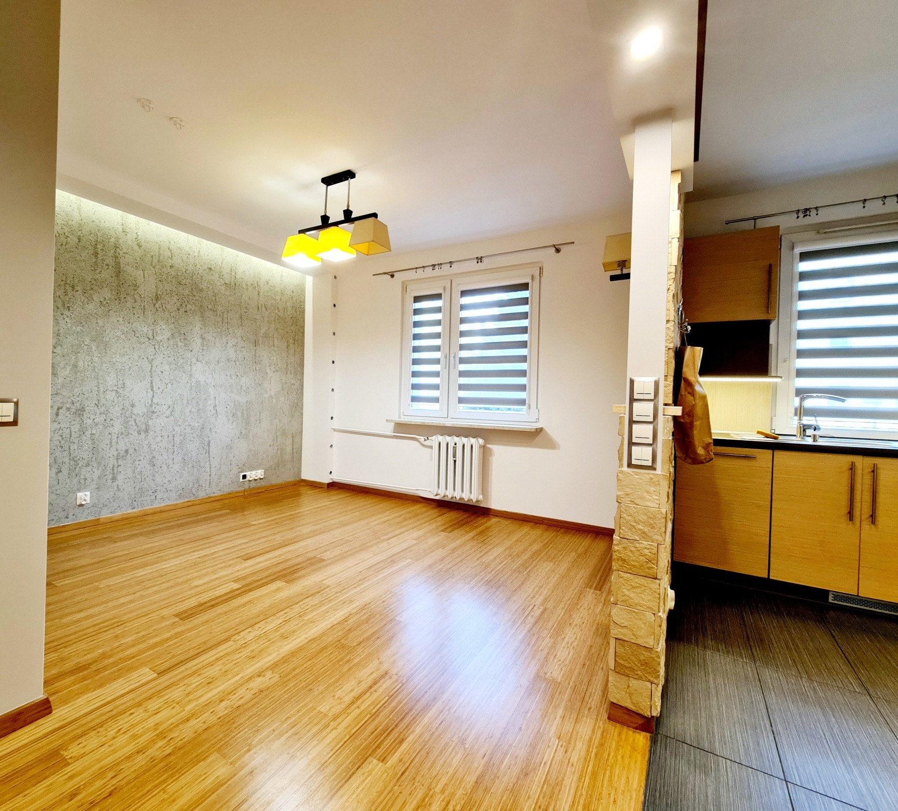 Puste, jasne mieszkanie z drewnianą podłogą, szarą ścianą akcentującą, żółtymi lampami i częściowo widoczną kuchnią z drewnianymi szafkami.