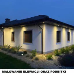 Oświetlona elewacja domu jednorodzinnego z ciemnymi oknami i dachem w wieczornym oświetleniu, widoczne podbitki i oświetlenie ogrodowe.