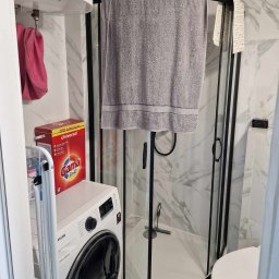 PROBUILD - Usługi ogólnobudowlane - Wykończona łazienka z kabiną prysznicową z czarnymi ramami, pralką Samsung, podgrzewaczem wody Ariston i marmurowymi płytkami na ścianach.