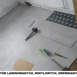 PROBUILD - Usługi ogólnobudowlane - Układanie szarych paneli podłogowych w pomieszczeniu, widoczne narzędzia: gumowy młotek, szczotka, dłuto, fragmenty podkładu i niedokończona powierzchnia obok kratki wentylacyjnej.