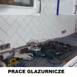 Montaż białych, prostokątnych płytek ułożonych w jodełkę na ścianie kuchni, widoczne gniazdka elektryczne, poziomica laserowa, narzędzia glazurnicze, kuchenka gazowa i białe szafki kuchenne.