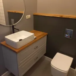 Nowoczesna łazienka z umywalką nablatową w kształcie prostokąta z czarnym obramowaniem, okrągłe lustro, drewniany blat i heksagonalne płytki podłogowe w odcieniach szarości.