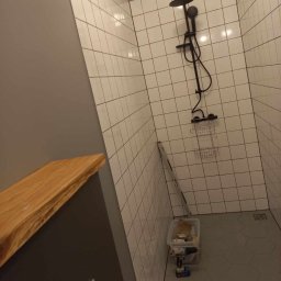 PROBUILD - Usługi ogólnobudowlane - Nowoczesna łazienka z białymi kwadratowymi płytkami na ścianach i heksagonalnymi szarymi płytkami na podłodze, czarna armatura prysznicowa, drewniany blat, drabina oparta o ścianę i pojemnik...