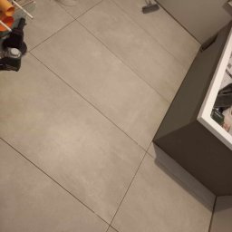 PROBUILD - Usługi ogólnobudowlane - Ułożona podłoga z jasnych, kwadratowych płytek o wymiarach 120 cm, widoczne narzędzia glazurnicze, pędzle, wiadro po farbie, szczotka i elementy instalacji kanalizacyjnej w rogu łazienki.