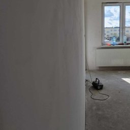 PROBUILD - Usługi ogólnobudowlane - Wnętrze w trakcie prac wykończeniowych: gładź na ścianie, szlifierka pod ścianą, widok na okno i grzejnik w tle.