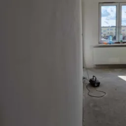 Wnętrze w trakcie prac wykończeniowych: gładź na ścianie, szlifierka pod ścianą, widok na okno i grzejnik w tle.