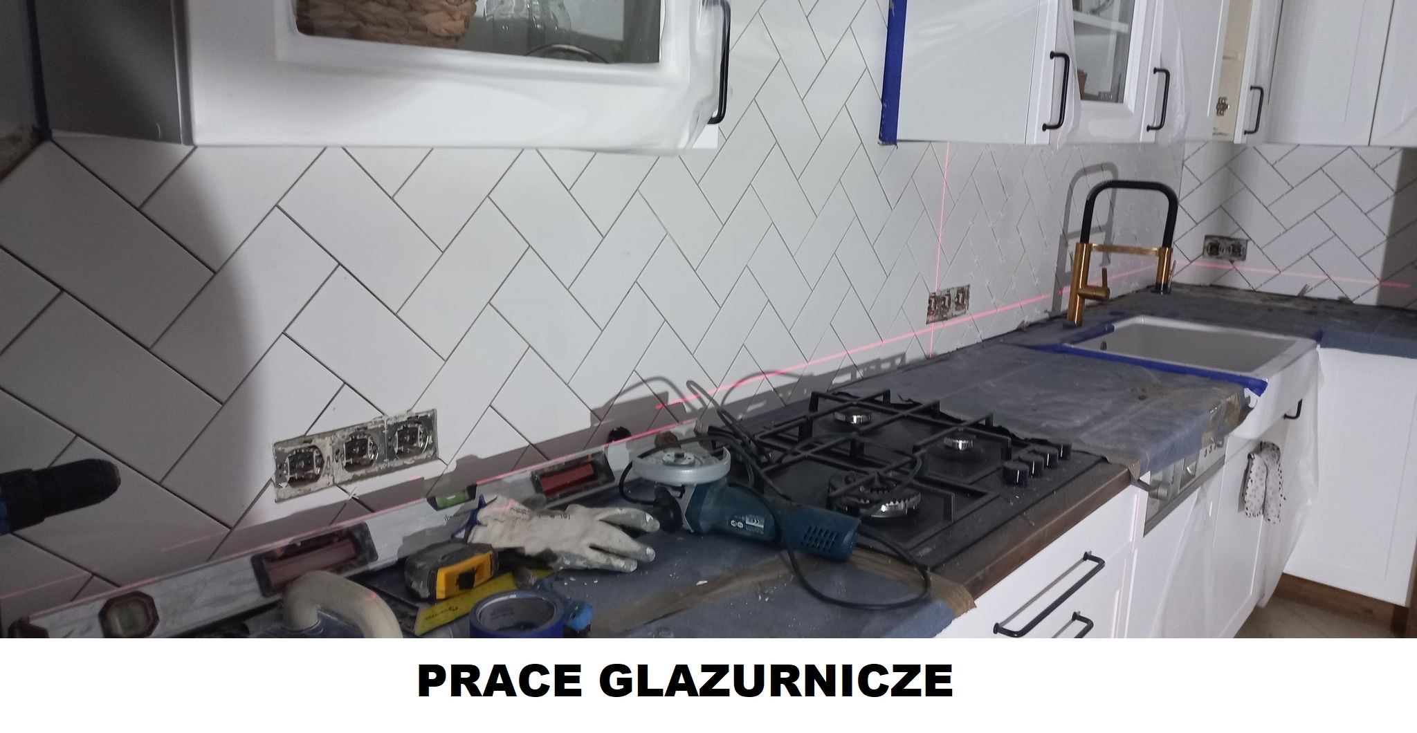 Montaż białych, prostokątnych płytek ułożonych w jodełkę na ścianie kuchni, widoczne gniazdka elektryczne, poziomica laserowa, narzędzia glazurnicze, kuchenka gazowa i białe szafki kuchenne.