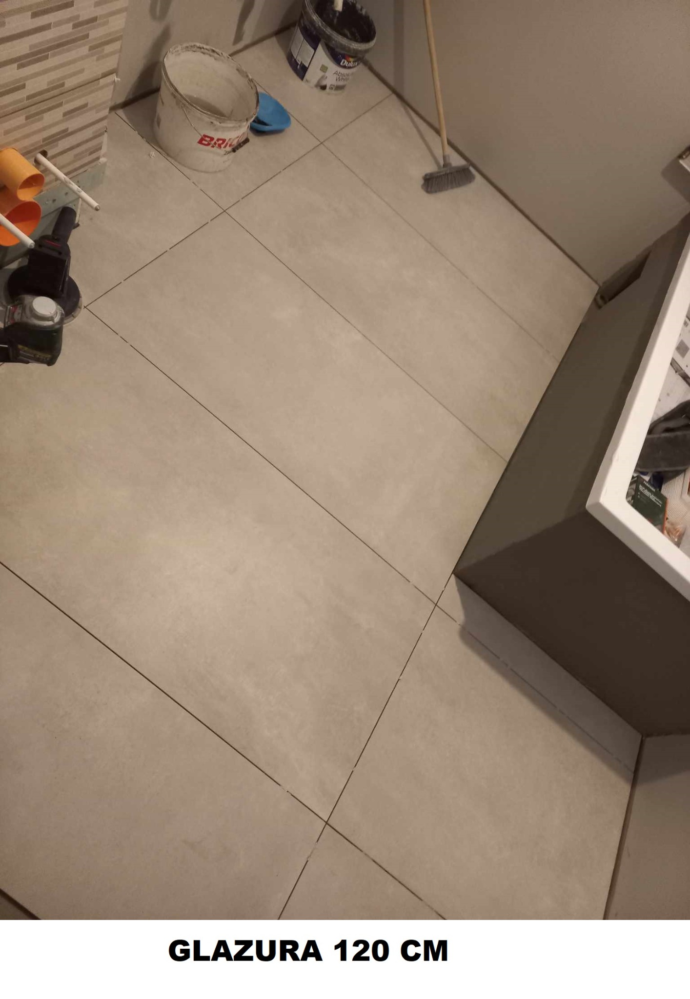 Ułożona podłoga z jasnych, kwadratowych płytek o wymiarach 120 cm, widoczne narzędzia glazurnicze, pędzle, wiadro po farbie, szczotka i elementy instalacji kanalizacyjnej w rogu łazienki.