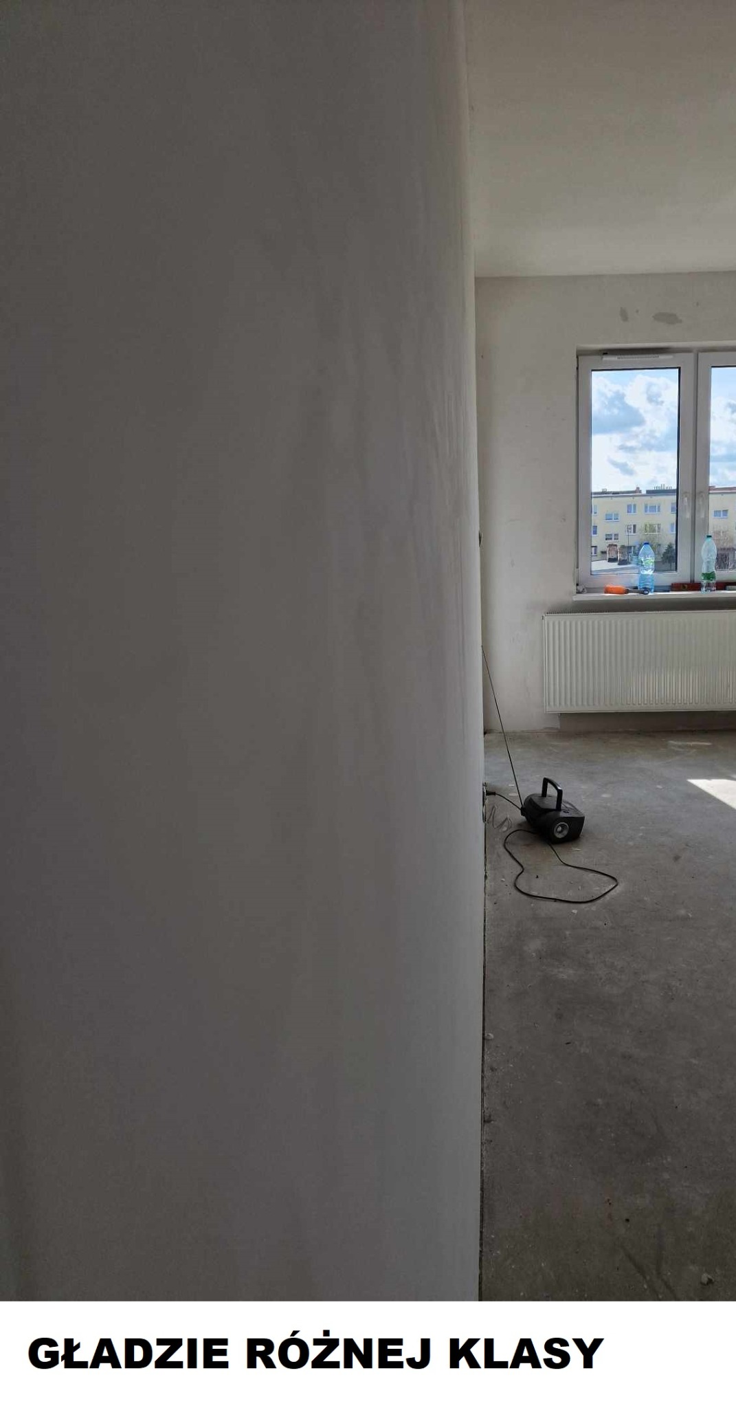 Wnętrze w trakcie prac wykończeniowych: gładź na ścianie, szlifierka pod ścianą, widok na okno i grzejnik w tle.