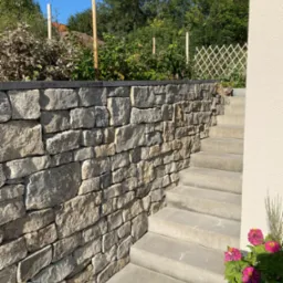 Murowana ściana oporowa z kamienia naturalnego, przylegająca do betonowych schodów prowadzących w górę, z roślinnością w tle i kwiatami w donicy w prawym dolnym rogu.