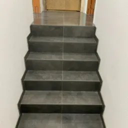 Schody wyłożone ciemnoszarą płytką, widoczne fragmenty niedokończonych prac wykończeniowych przy podłodze i ścianach.