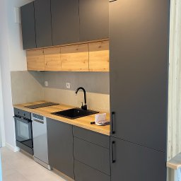 Usługi Glazurnicze MAGNAK - Nowoczesna kuchnia z szarymi szafkami i drewnianymi akcentami. Czarny zlew i bateria, jasny blat. Minimalistyczny design, widok na całą zabudowę kuchenną.