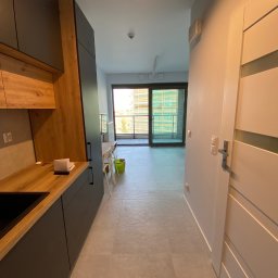 Usługi Glazurnicze MAGNAK - Nowoczesna kuchnia z drewnianymi akcentami i szarymi szafkami, widok na jasny salon z balkonem. Minimalistyczny design, świeże wykończenie, płytki podłogowe w odcieniach szarości.