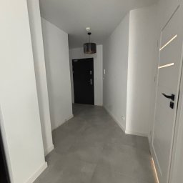 Usługi Glazurnicze MAGNAK - Nowoczesny korytarz z szarymi płytkami podłogowymi, białymi ścianami i czarnymi drzwiami wejściowymi. Minimalistyczna lampa sufitowa i białe drzwi wewnętrzne z czarną klamką.