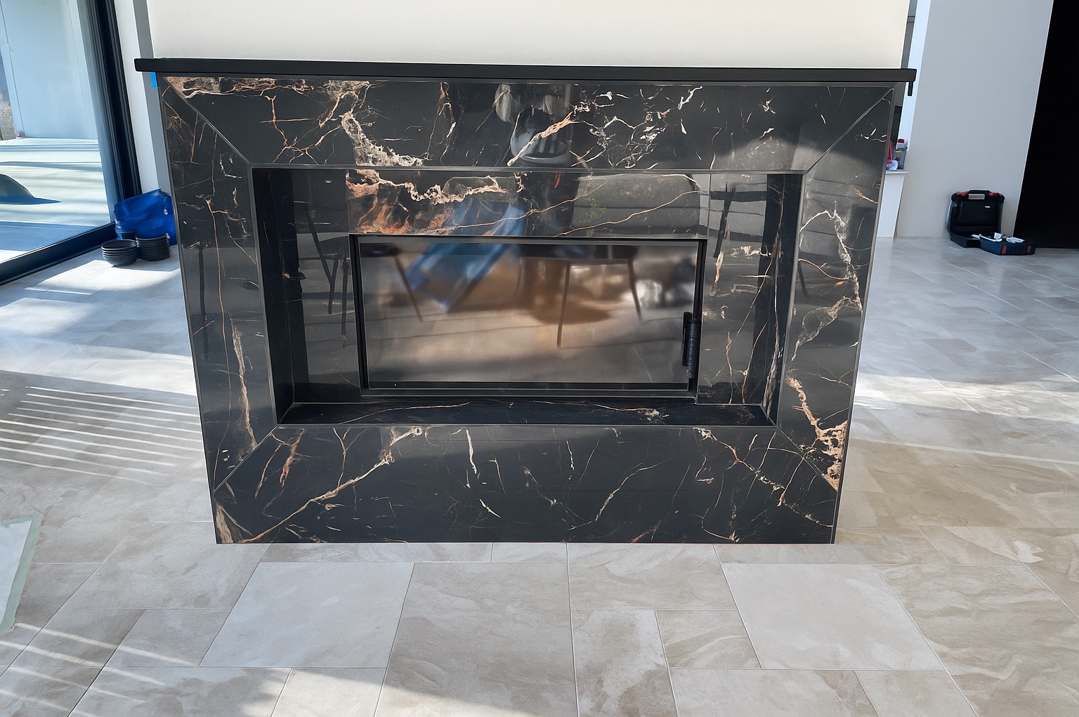 Obudowa kominka z czarnego marmuru z wyraźnym, złotym żyłkowaniem, dopasowana do podłogi z płytek w odcieniach beżu i szarości. Nowoczesny design.