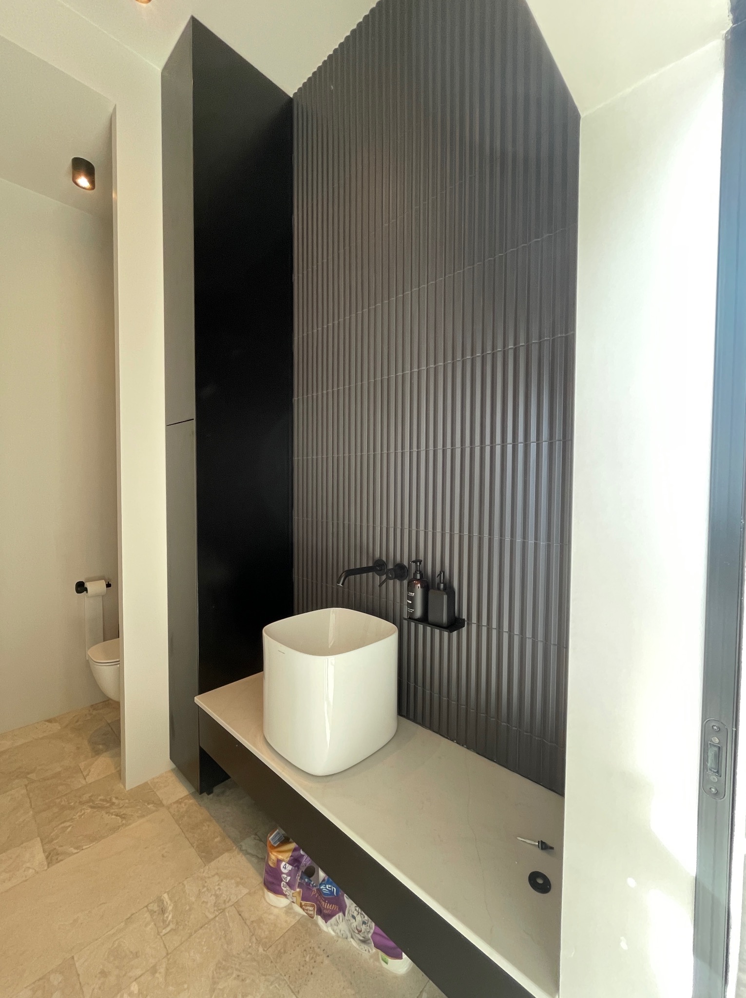 Nowoczesna łazienka z umywalką nablatową, czarną armaturą i płytkami w pionowe prążki. Widoczny fragment toalety i podłoga z jasnego kamienia.