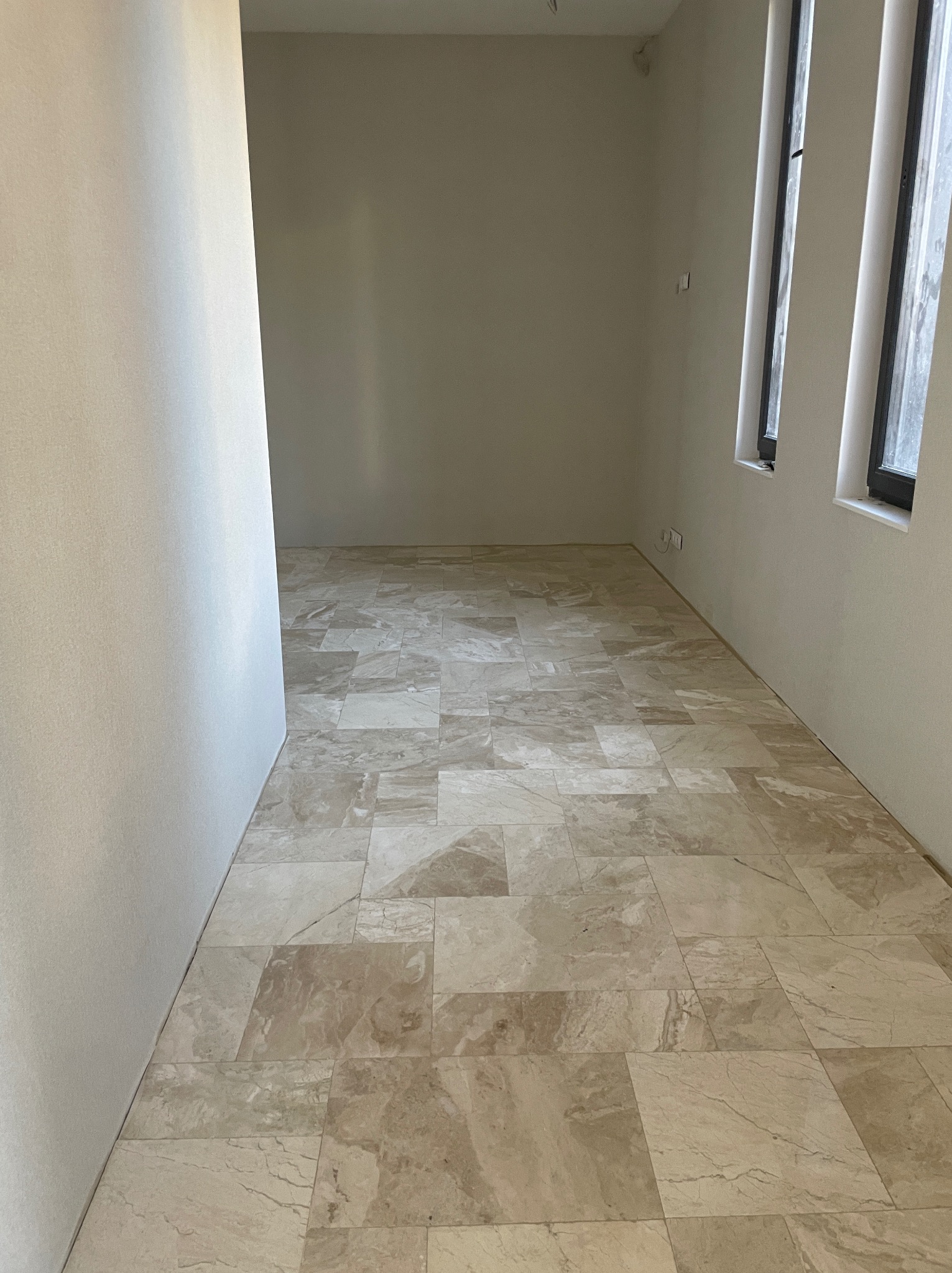Jasna, marmurkowa podłoga z płytek w pustym pokoju z oknami. Ściany pomalowane na kremowo. Widok na świeżo wykończone wnętrze.