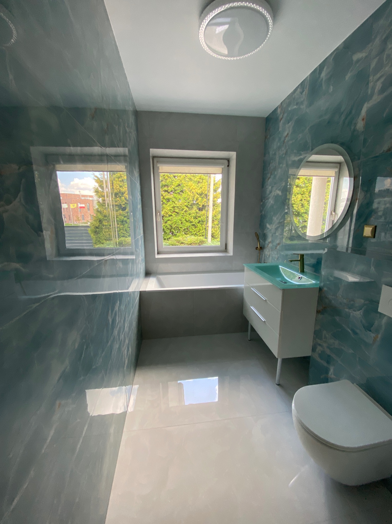 Wykończona łazienka z turkusowymi płytkami imitującymi marmur, białą armaturą i oknem z widokiem na zieleń. Nowoczesny design z okrągłym lustrem i minimalistyczną lampą sufitową.