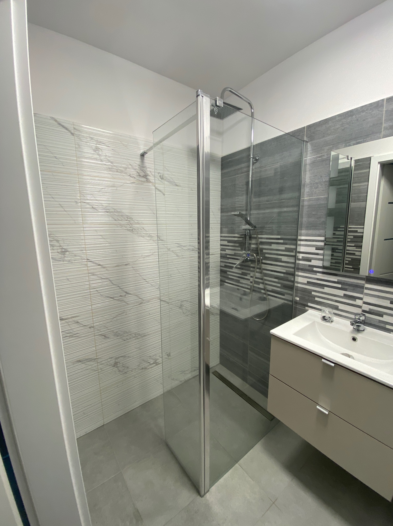 Nowoczesna łazienka po remoncie: kabina prysznicowa ze szklaną ścianką, umywalka z szafką w odcieniach beżu, płytki imitujące marmur i szare płytki w geometryczny wzór.