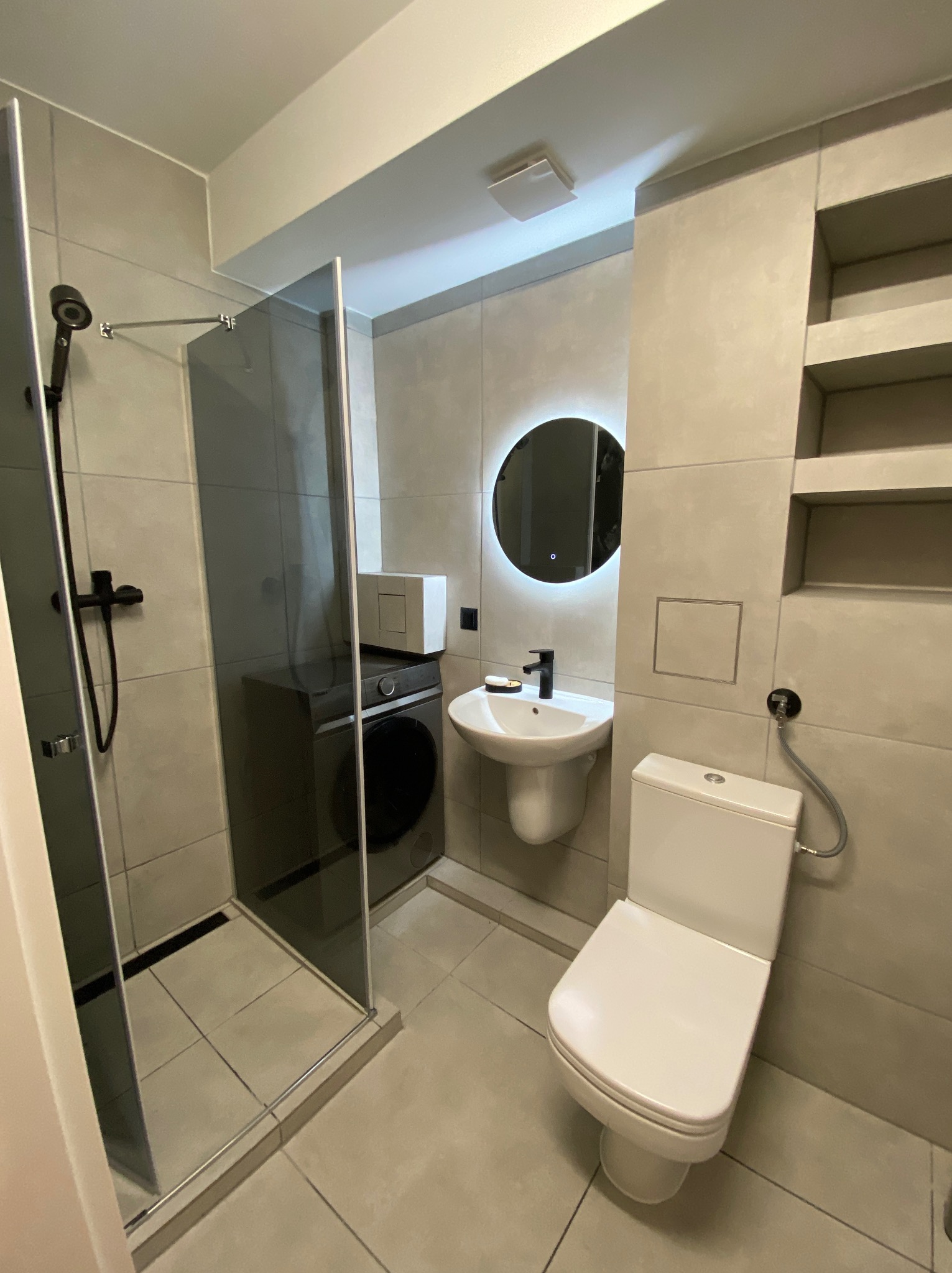 Nowoczesna łazienka w odcieniach szarości z kabiną prysznicową typu walk-in, podświetlanym lustrem i pralką w zabudowie. Minimalistyczny design i funkcjonalne rozwiązania.