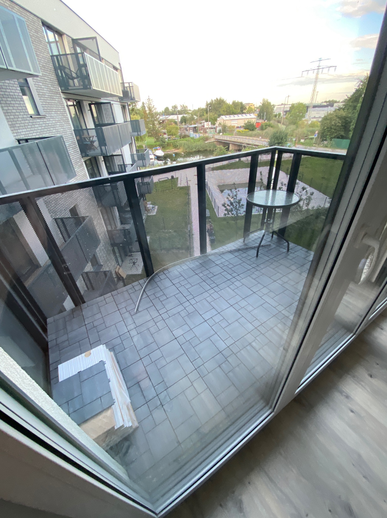 Wykończony balkon w bloku z szarymi płytkami, stolikiem i widokiem na zieleń. Na podłodze leżą zapakowane płytki. Widok z wnętrza mieszkania.