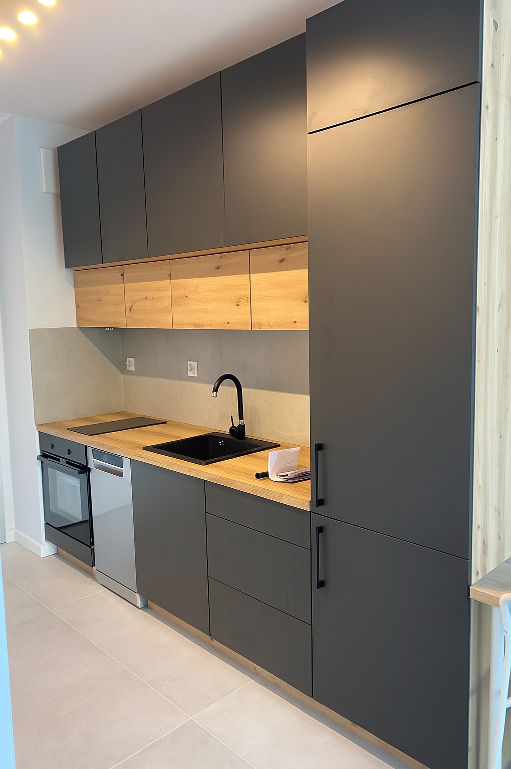 Nowoczesna kuchnia z szarymi szafkami i drewnianymi akcentami. Czarny zlew i bateria, jasny blat. Minimalistyczny design, widok na całą zabudowę kuchenną.