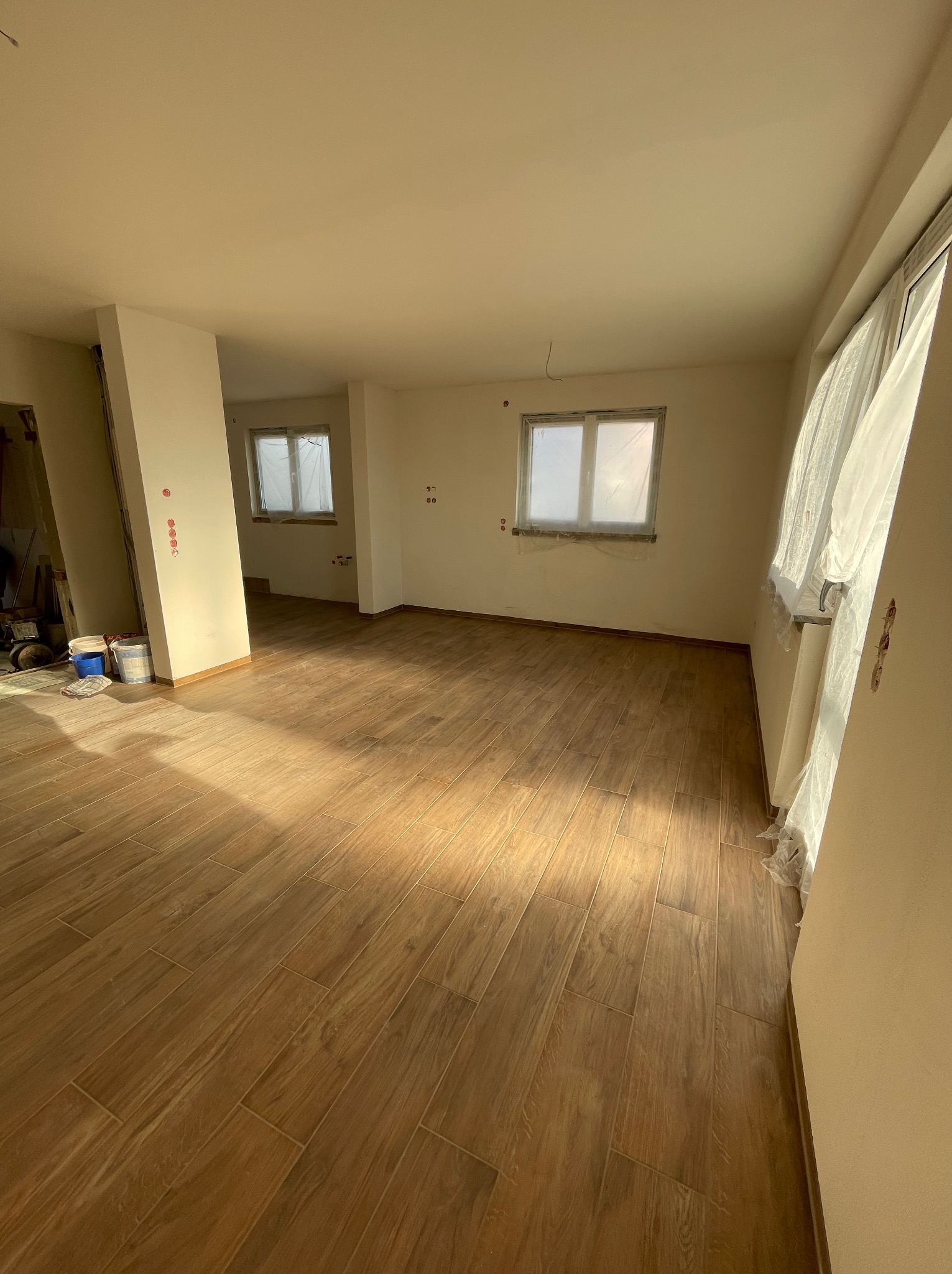 Jasne wnętrze po remoncie z drewnopodobną podłogą z płytek, zabezpieczone okna folią malarską, widoczne ślady po pracach wykończeniowych.