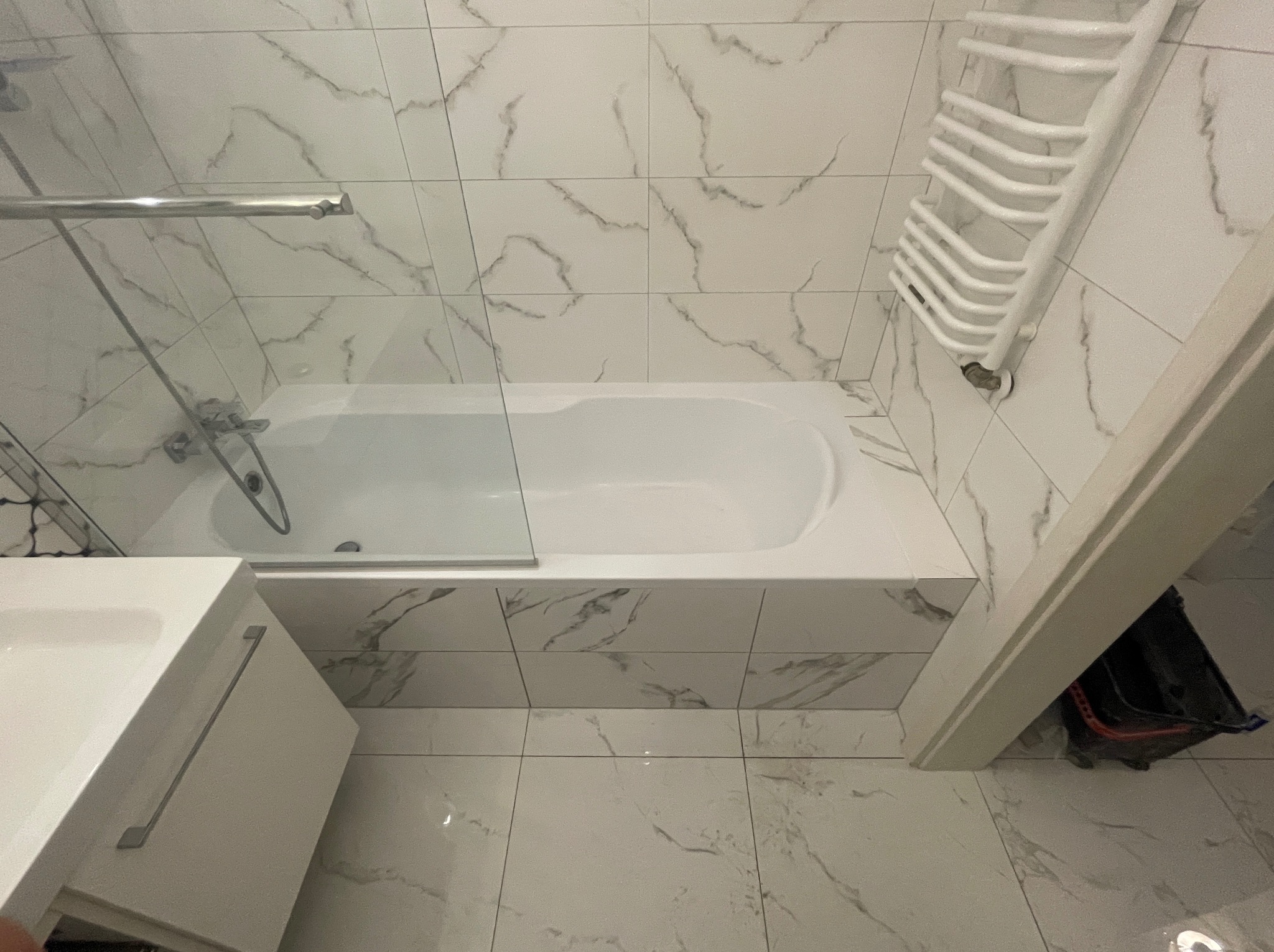 Biała wanna z kabiną prysznicową, umywalka z szafką i grzejnik drabinkowy na ścianie wyłożonej płytkami imitującymi marmur w wykończonej łazience.