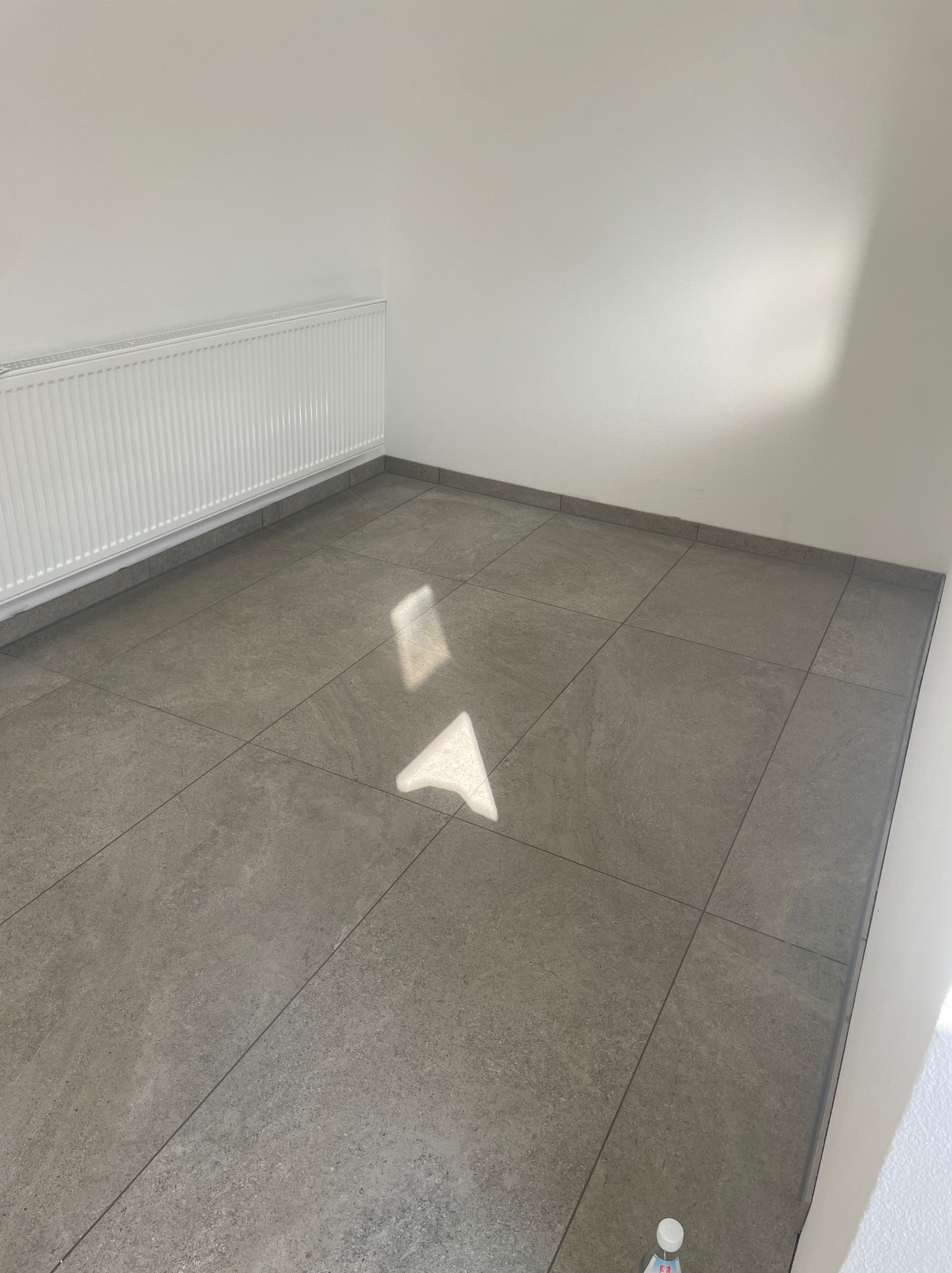 Wykończone wnętrze z szarymi płytkami podłogowymi i białym grzejnikiem. Widoczne delikatne refleksy światła na podłodze. Minimalistyczny styl.